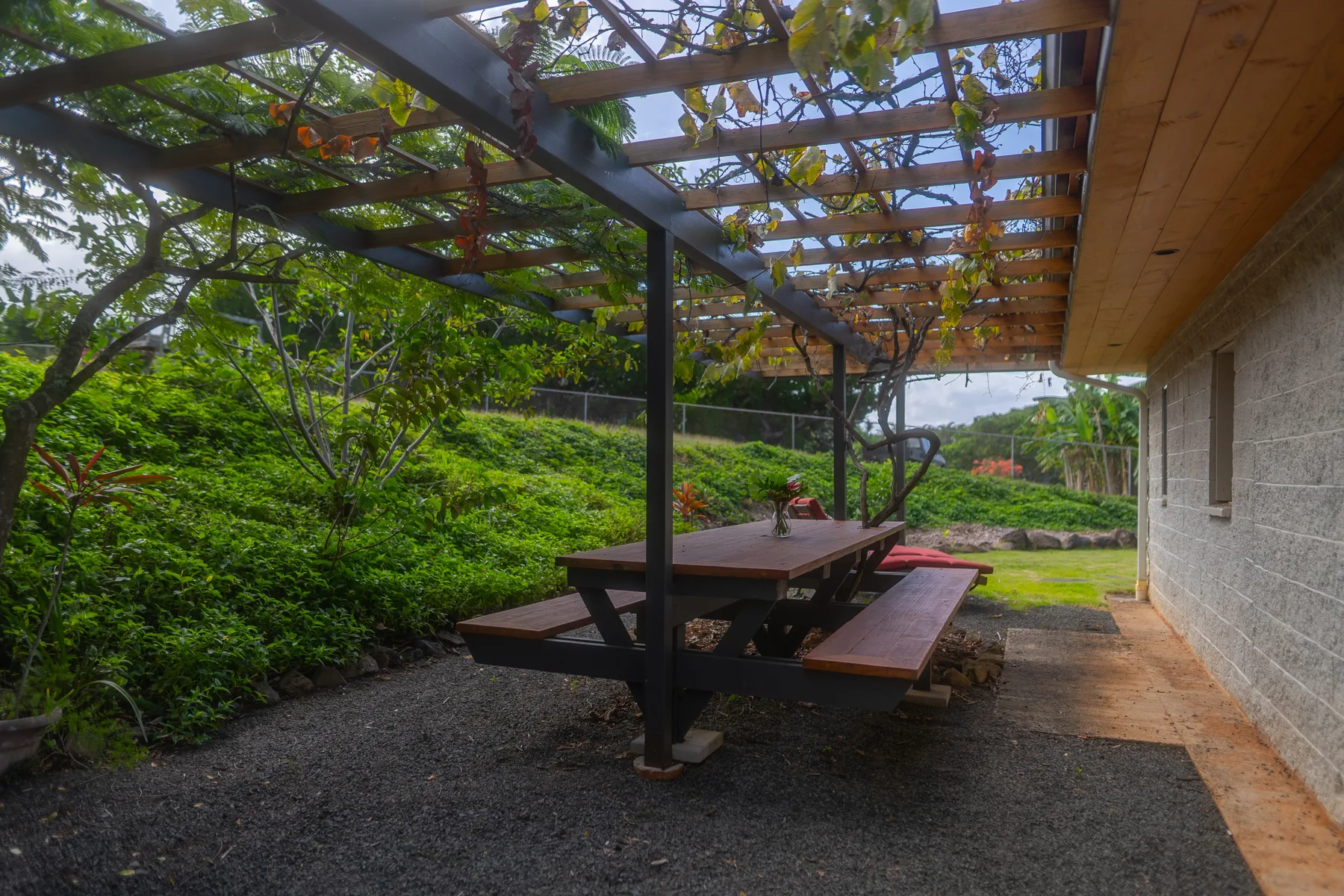 Una mesa de picnic está bajo una pérgola cubierta de enredaderas junto a un jardín.