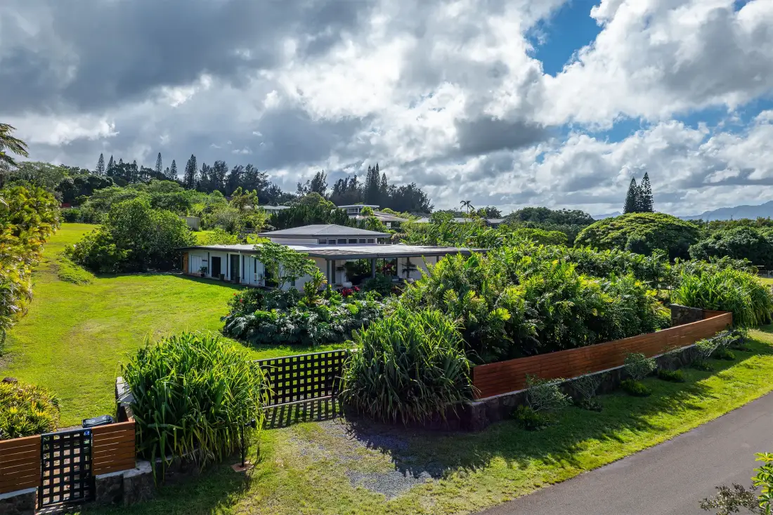 Una casa baja está rodeada de jardines frondosos junto a una carretera pavimentada y un cielo nublado.