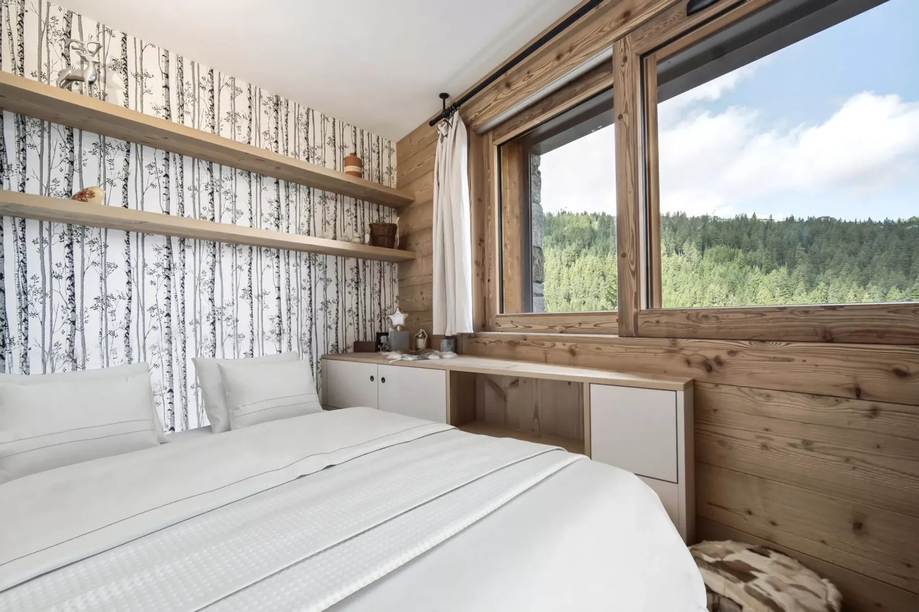 A bedroom with a neatly made bed beside a large window overlooking trees.