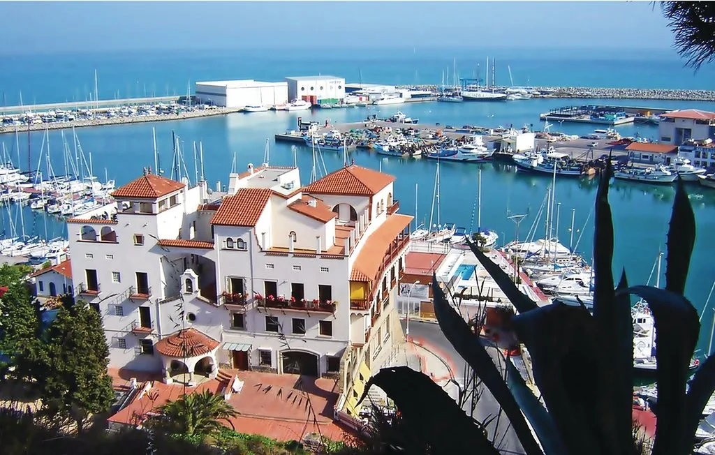 Una marina llena de barcos rodea un edificio blanco junto al agua con tejados de terracota.