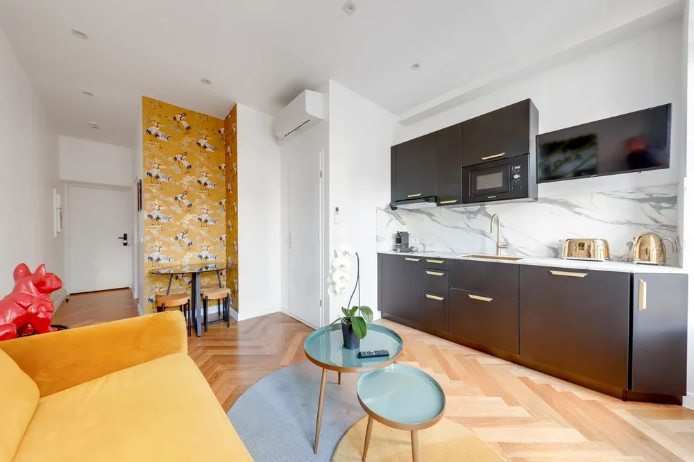 Una sala luminosa se abre a una cocina moderna, con un sofá amarillo y una pared estampada.