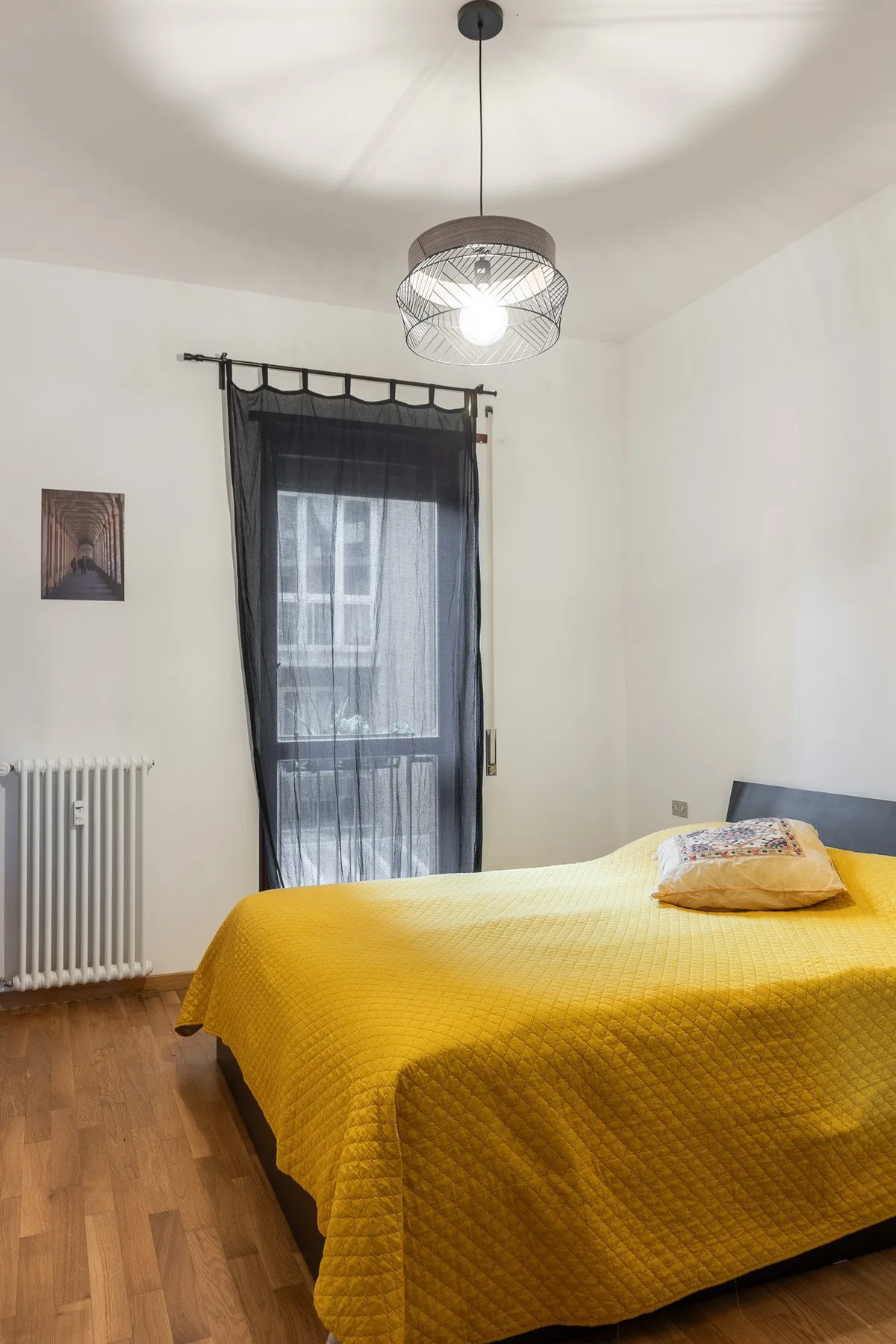 A bedroom with a bed covered in a yellow quilt beside a tall curtained window.