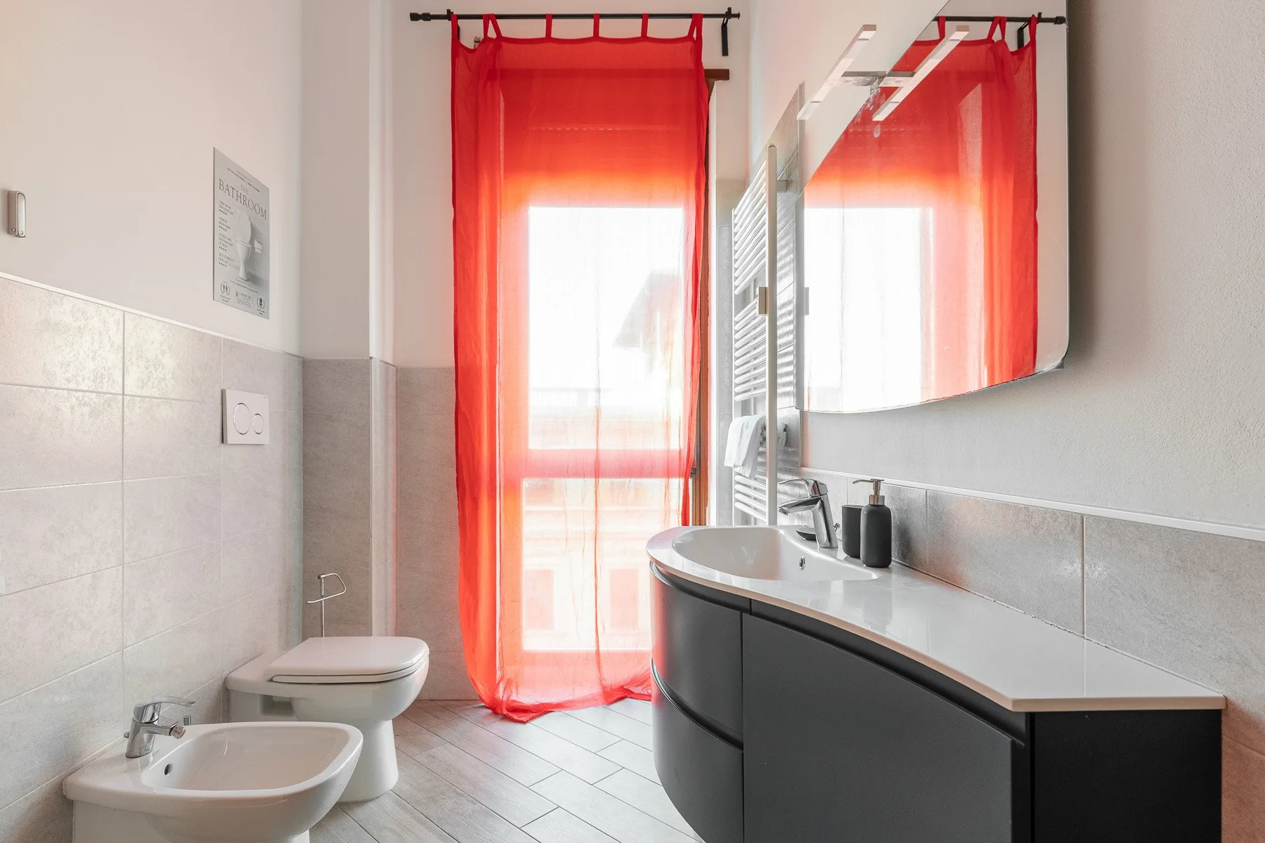 A tiled bathroom with a sink, toilet, and bright red curtains by the window.