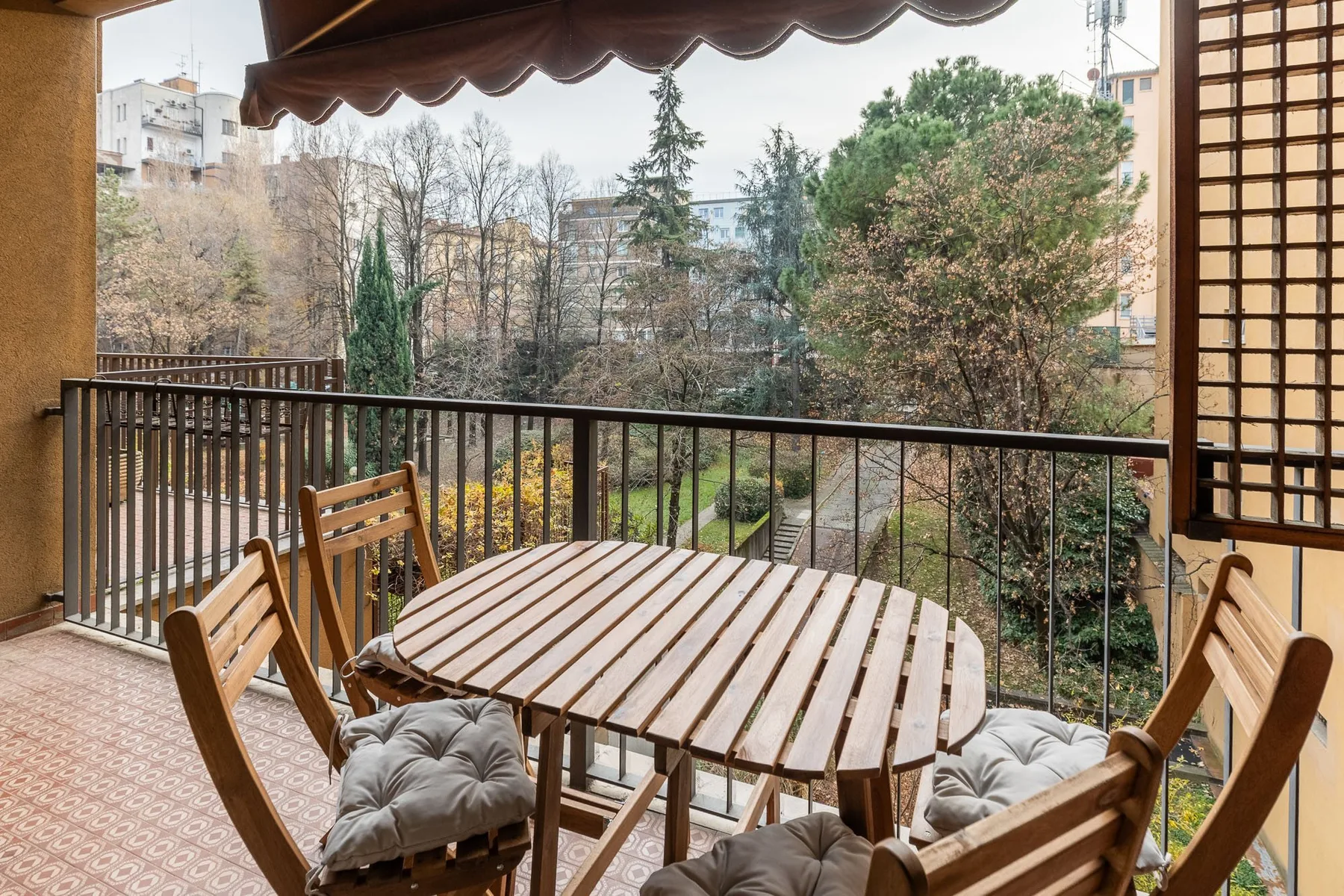 A balcony dining area with a wooden table and seating overlooks leafy trees.