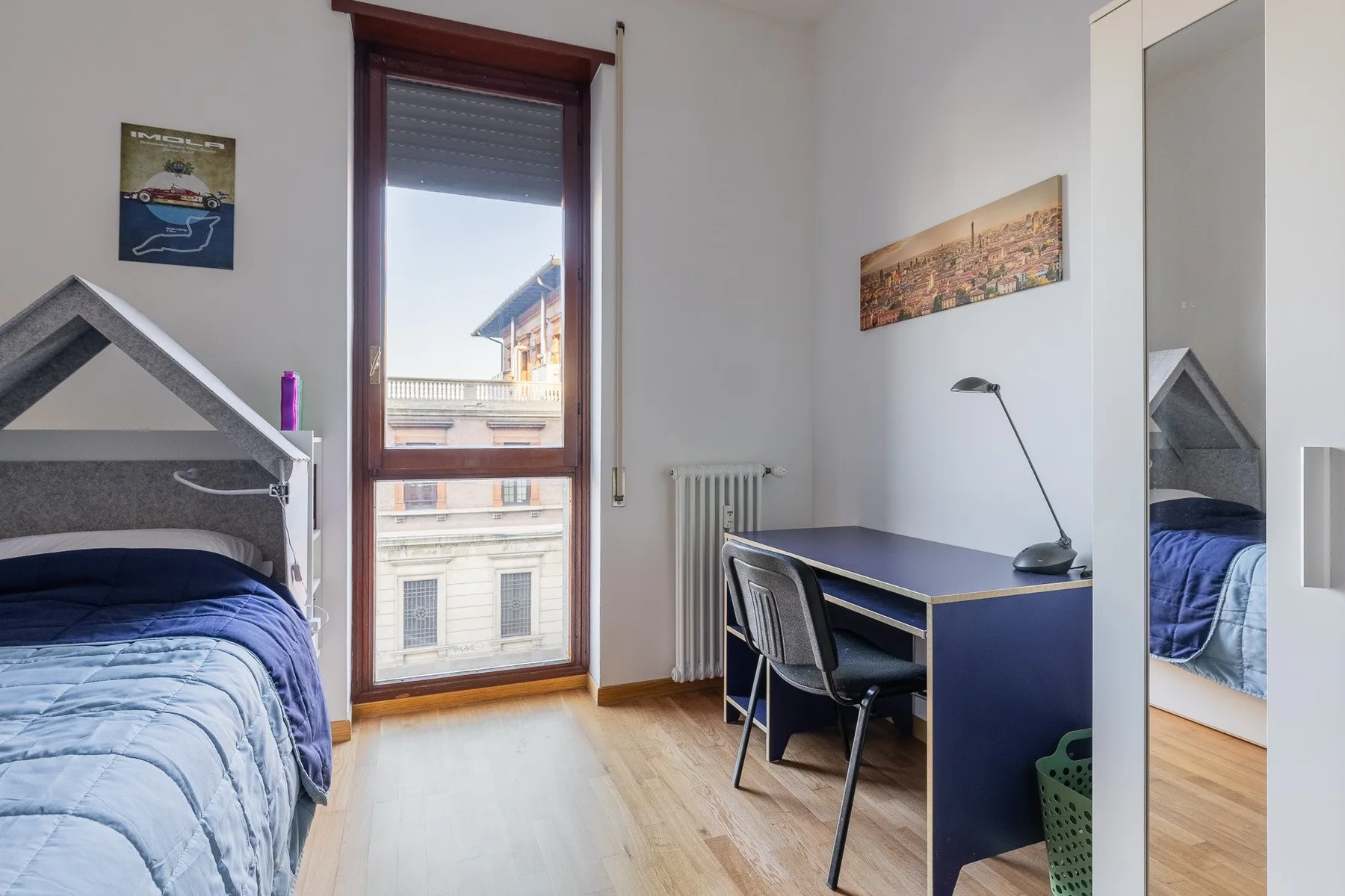 A bedroom with a desk and chair beside a tall window.
