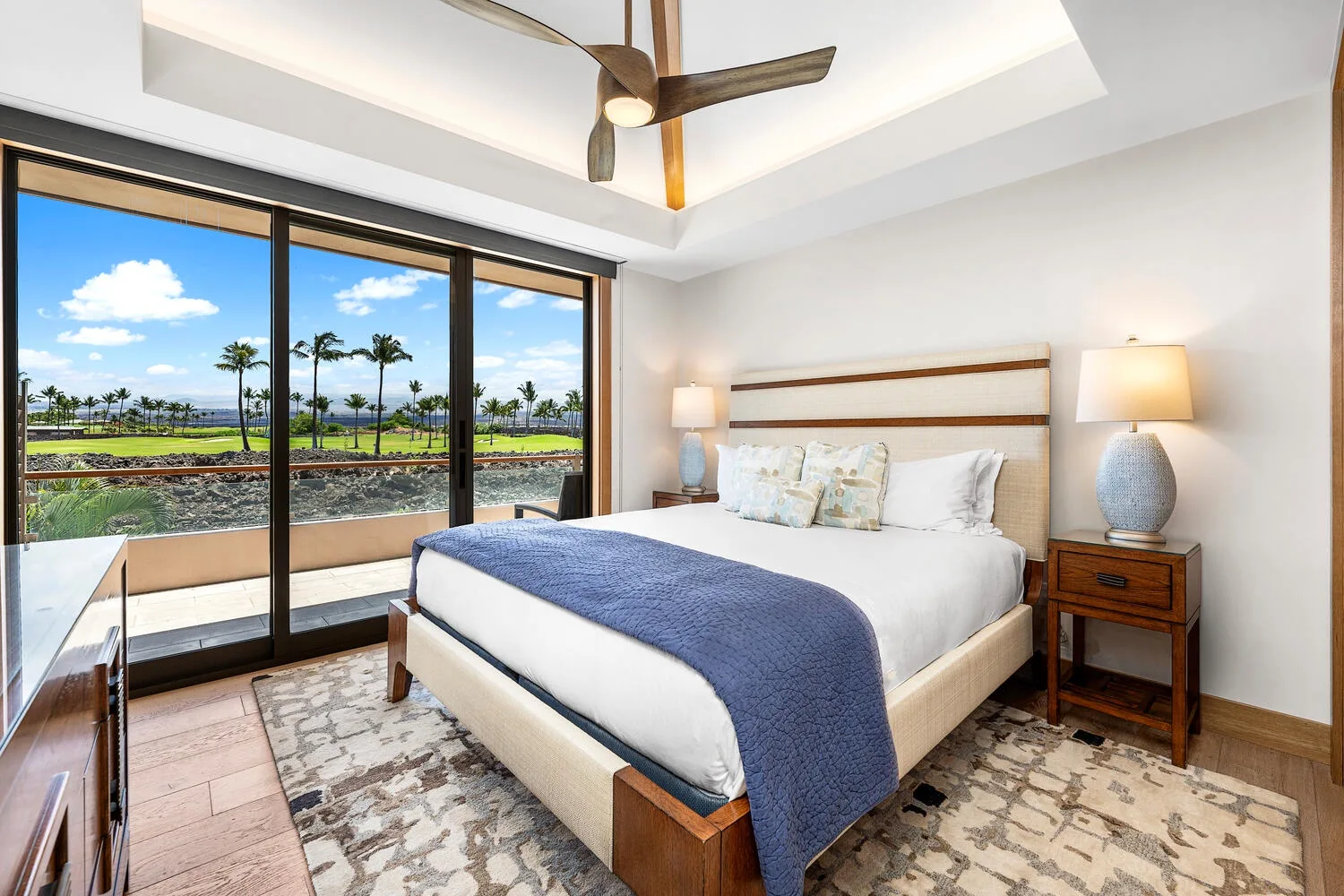 Un dormitorio luminoso con una cama tendida y grandes ventanas con vistas a palmeras.