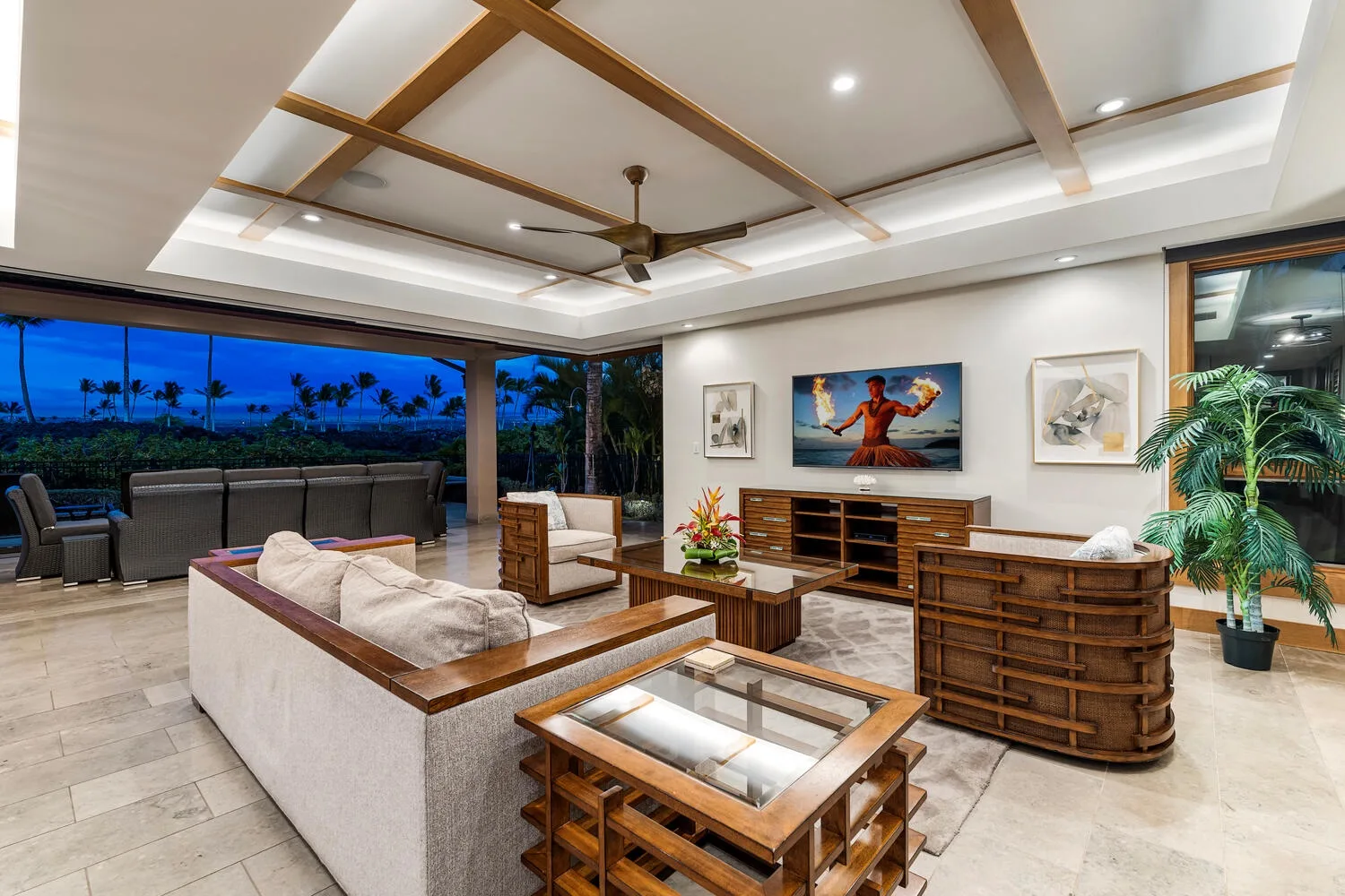 Una sala de estar amplia con asientos, muebles de madera y grandes ventanales con palmeras.