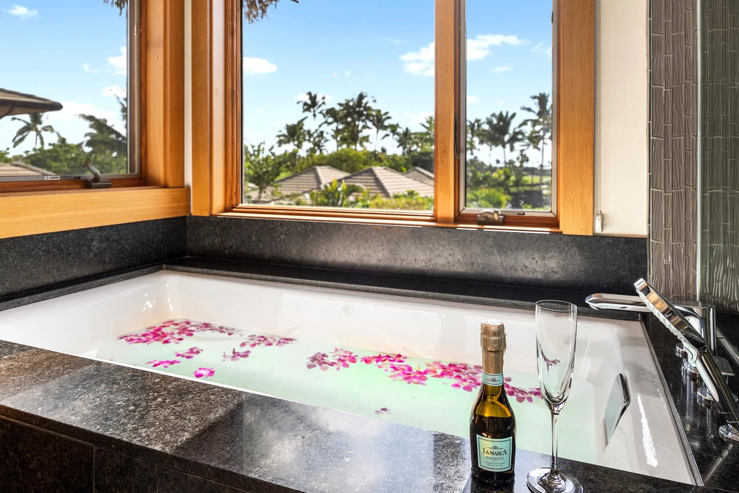 Una bañera con agua y pétalos de flores está junto a grandes ventanas.
