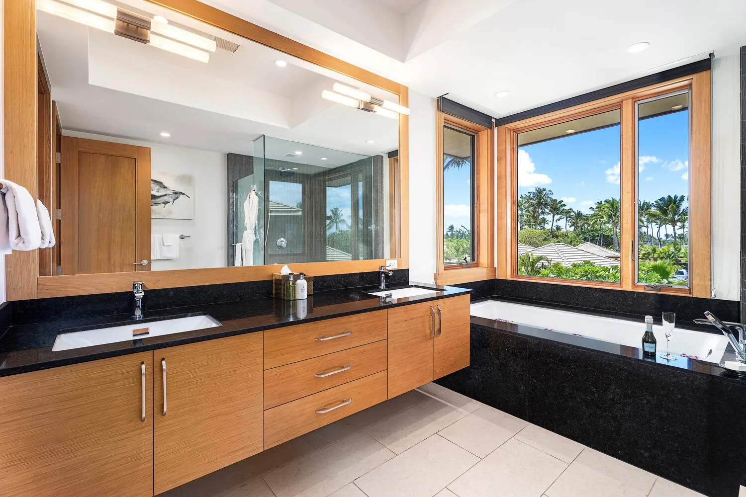 Un baño luminoso tiene un largo tocador de madera y grandes ventanas con vista a la vegetación.
