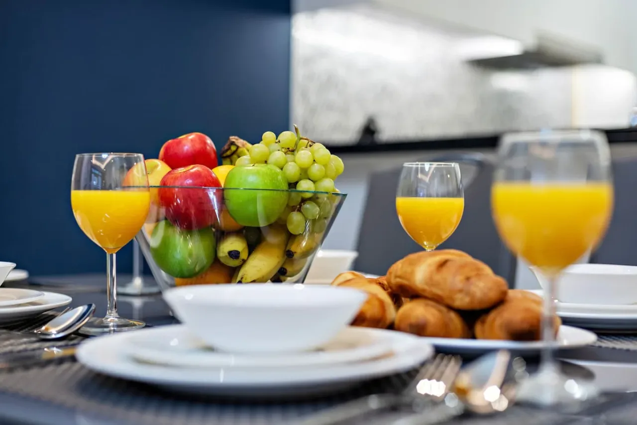 Una mesa está servida con fruta, bollería y vasos de zumo de naranja.