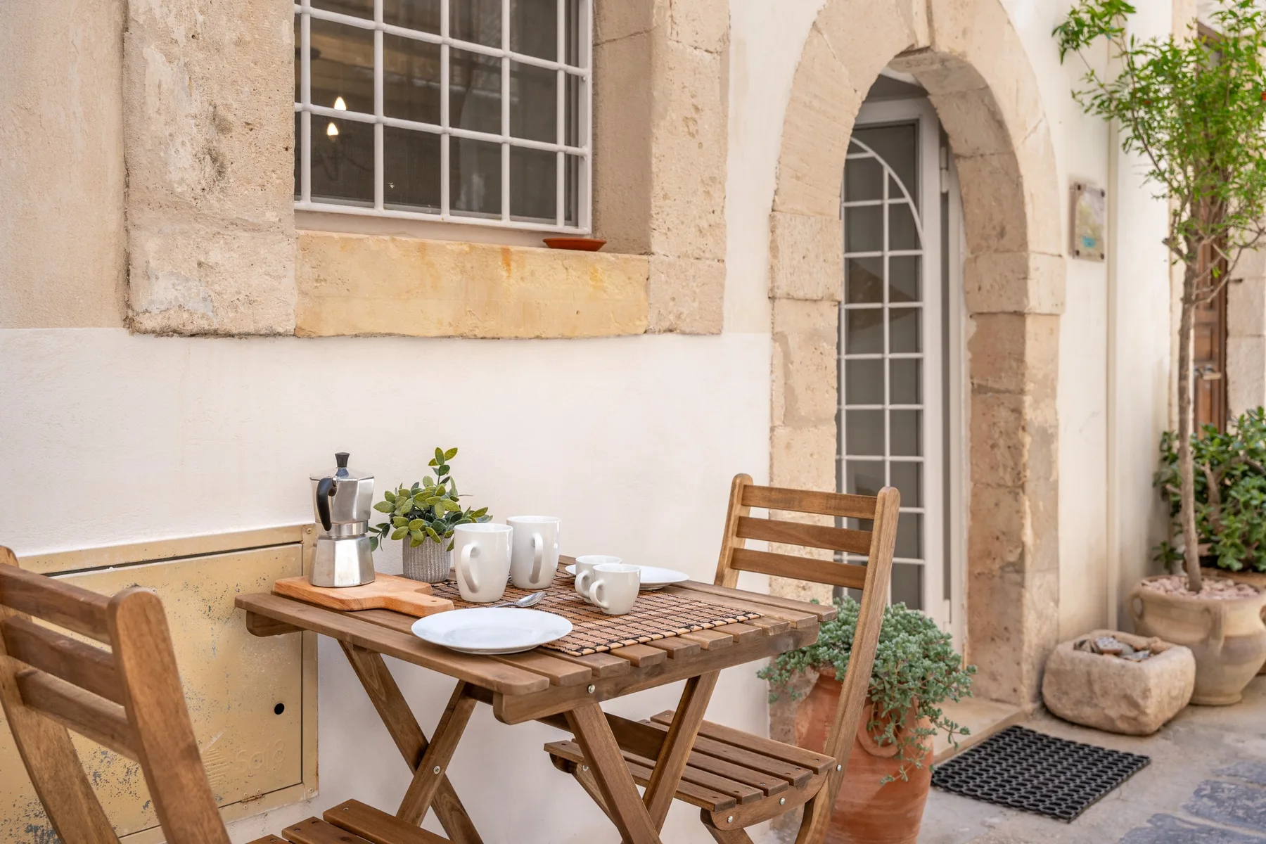Una pequeña zona de comedor al aire libre con una mesa de madera y tazas junto a una puerta arqueada.