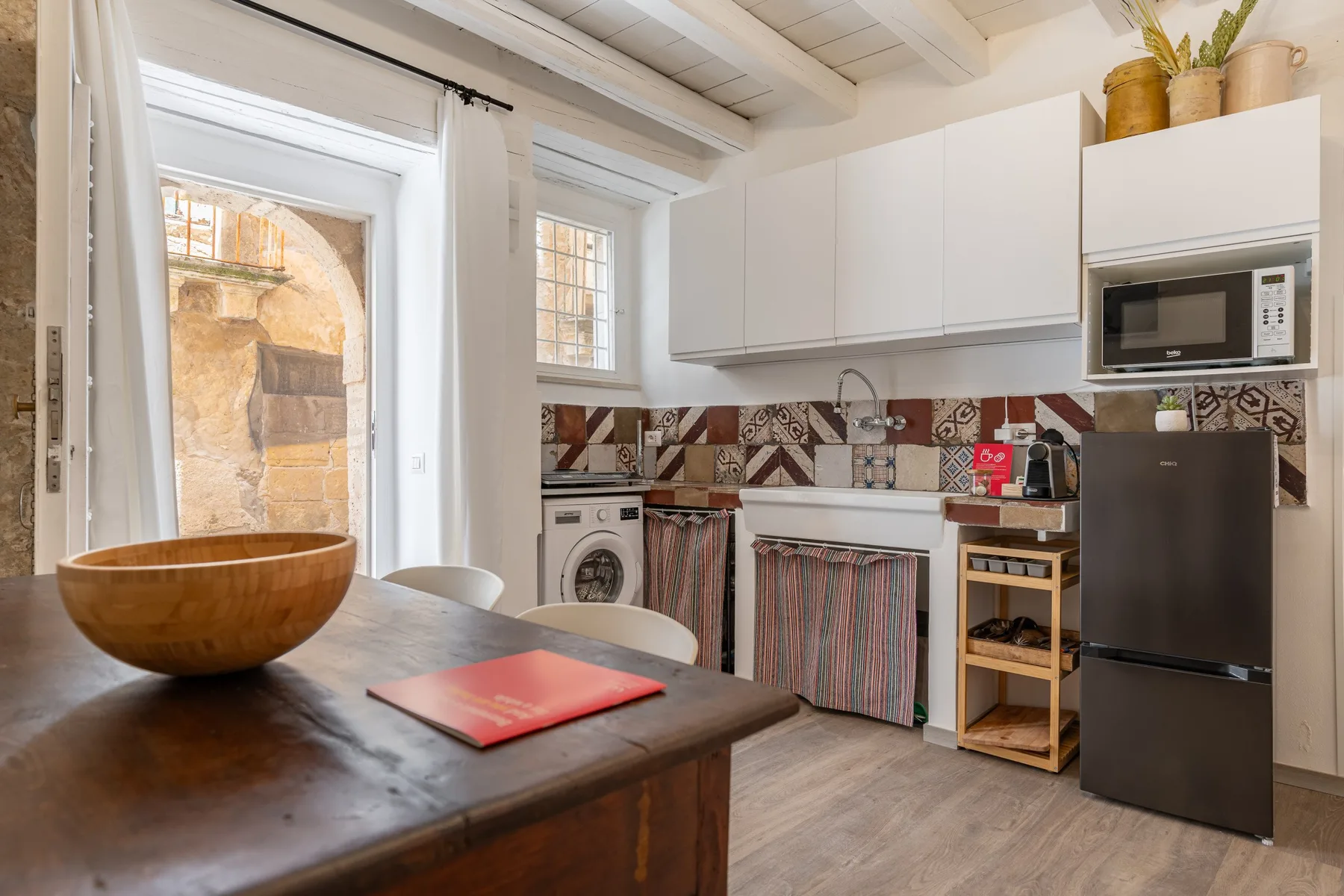 Una cocina luminosa con gabinetes blancos, un salpicadero de azulejos y una mesa de madera.