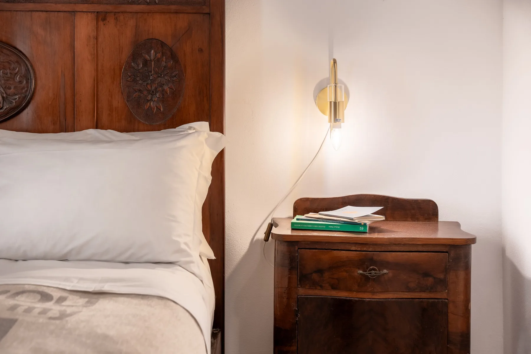 Un dormitorio con una cama junto a una mesita de madera y una luz de pared.