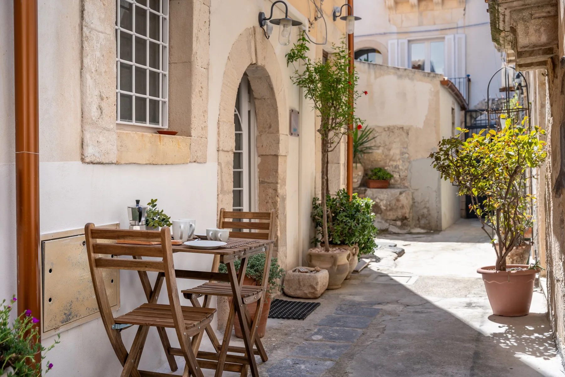 Un patio de piedra soleado tiene una mesa de madera y sillas junto a plantas en macetas.