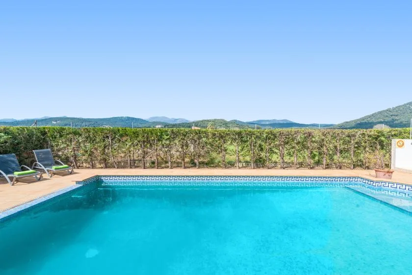 An outdoor swimming pool sits beside a hedge, with hills visible beyond.