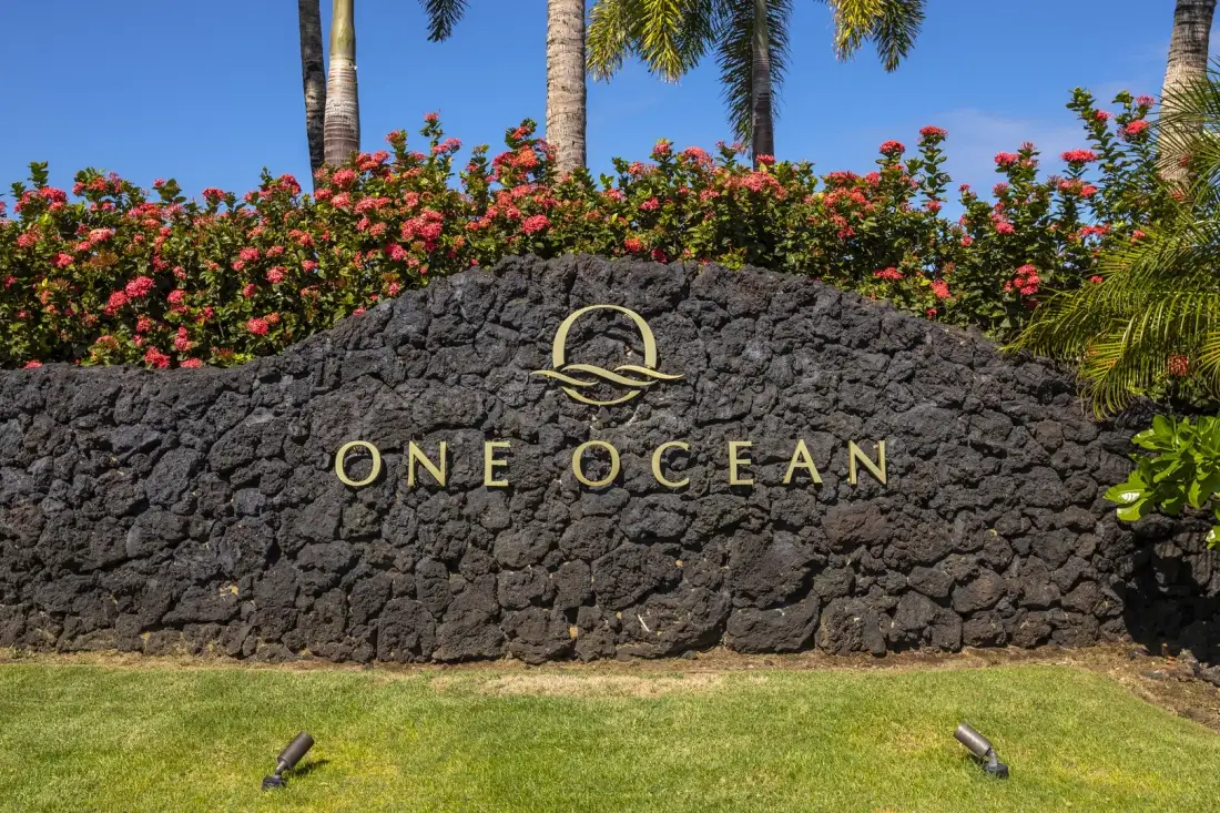 Un letrero de piedra ajardinado muestra las palabras «ONE OCEAN» bajo arbustos floridos.