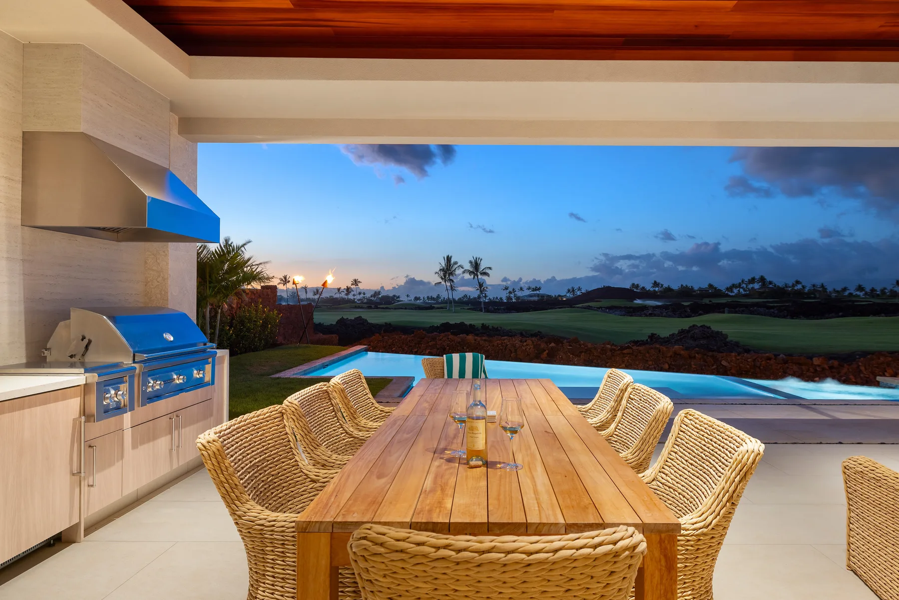Comedor al aire libre con mesa de madera y asientos de mimbre junto a una piscina.