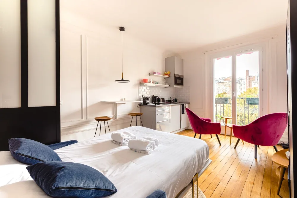 A bright studio bedroom with a bed, compact kitchen, and seating by the balcony doors.