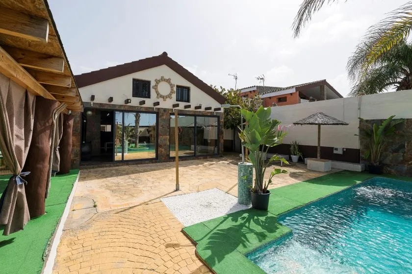 A swimming pool and paved patio sit beside a house with a covered outdoor seating area.