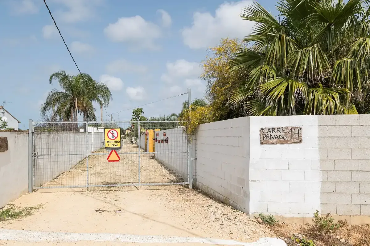 A gated sandy driveway runs between low walls and palm trees.