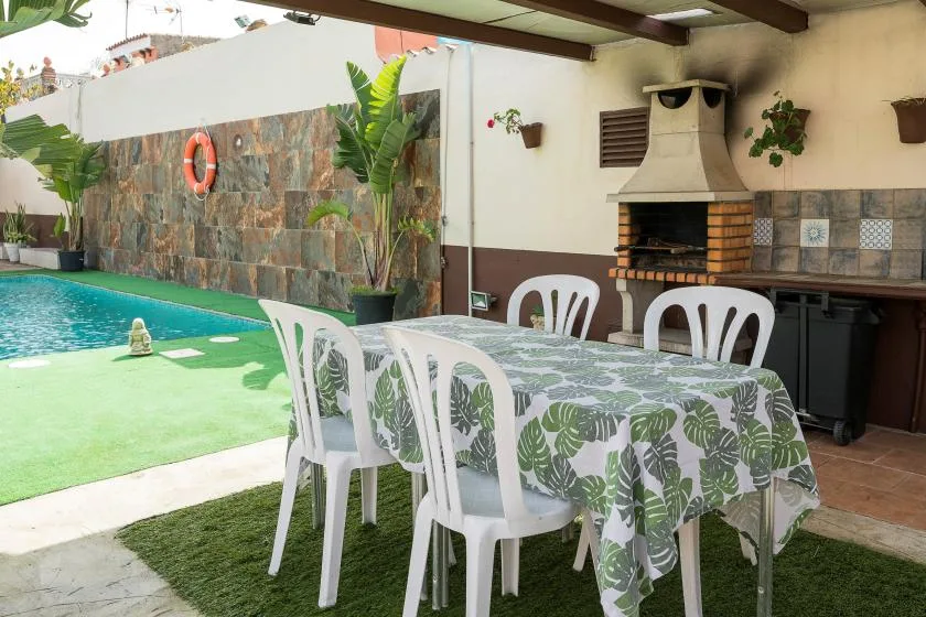 An outdoor dining area beside a swimming pool, with a covered grill in the background.