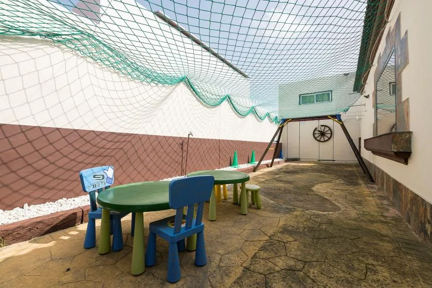 A paved outdoor play area has small tables and chairs beneath a protective net.