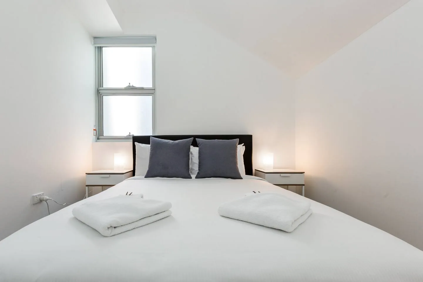 Un dormitorio luminoso tiene una cama bien tendida con almohadas oscuras y una ventana sobre el cabecero.