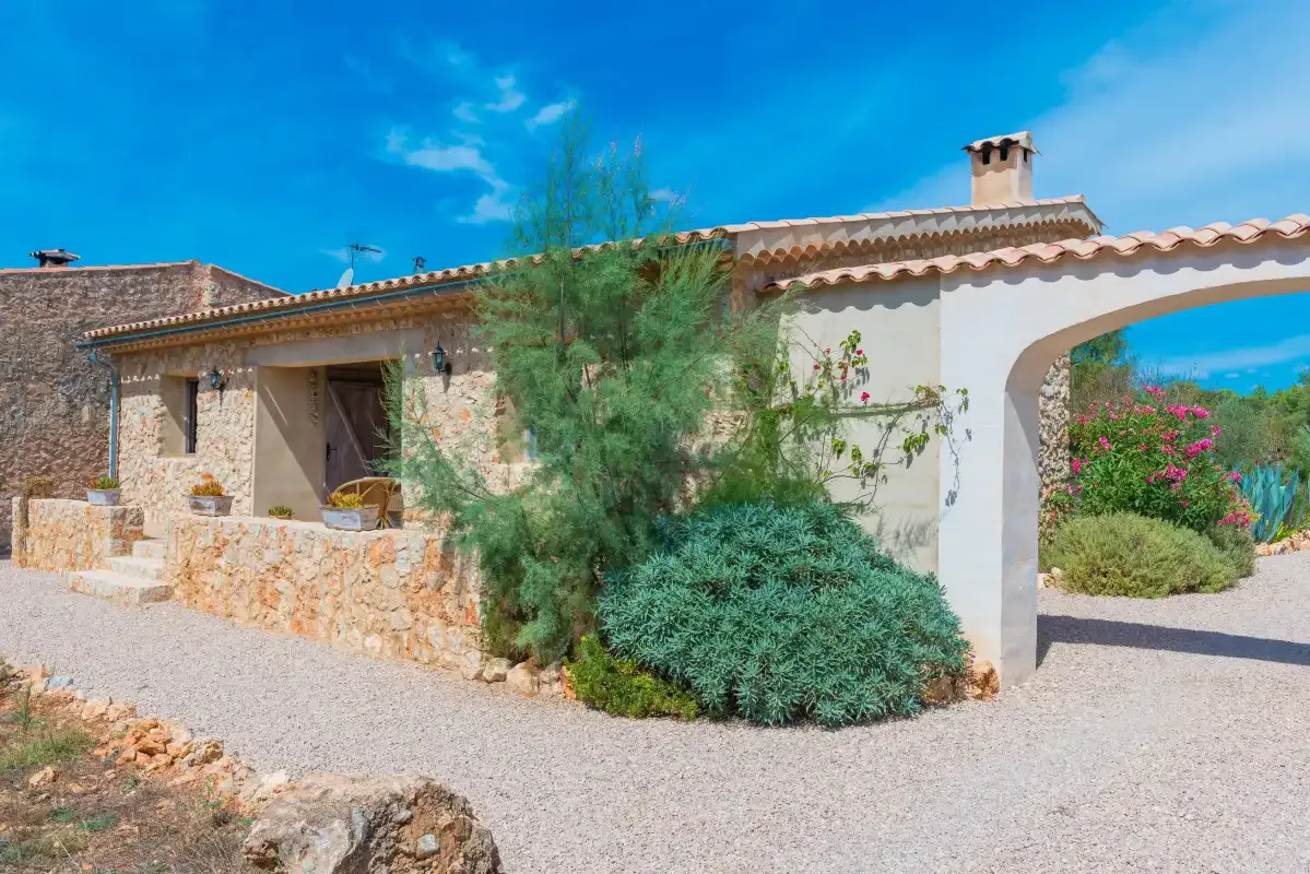 Una casa de piedra con techo de tejas está junto a un camino de grava y plantas con flores.