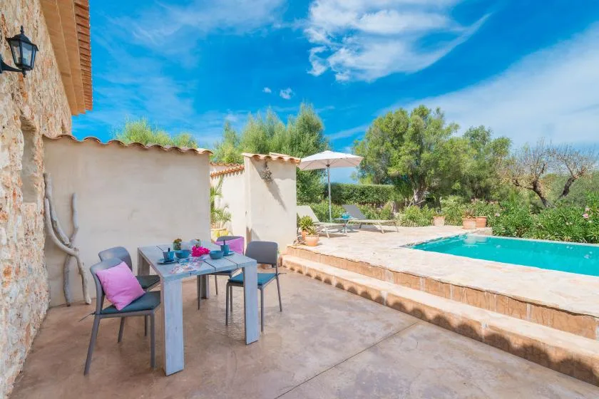 Una zona de comedor al aire libre junto a una piscina, con árboles y una sombrilla.