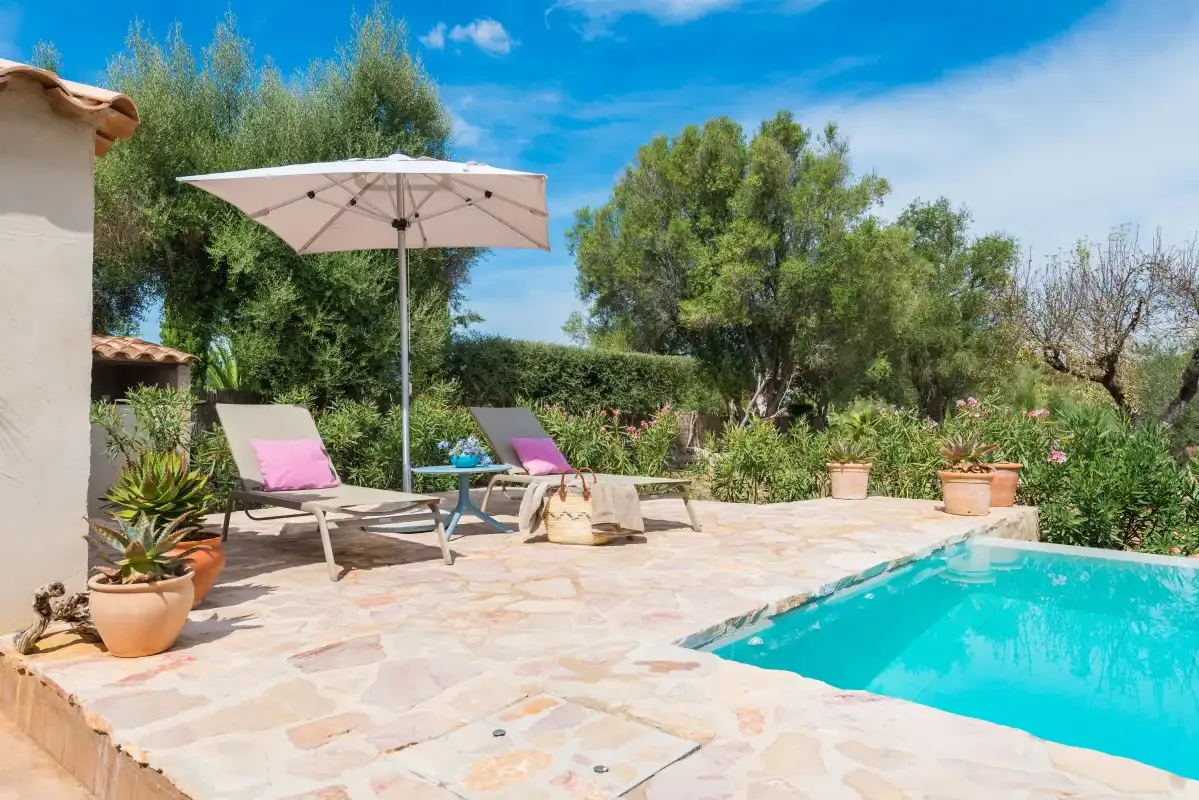 Un patio soleado con piscina, asientos, una sombrilla y vegetación alrededor.