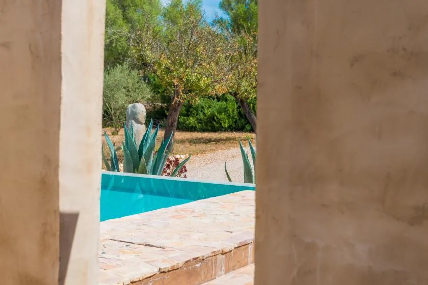 Una piscina y un patio de piedra quedan enmarcados por una puerta abierta, con árboles al fondo.