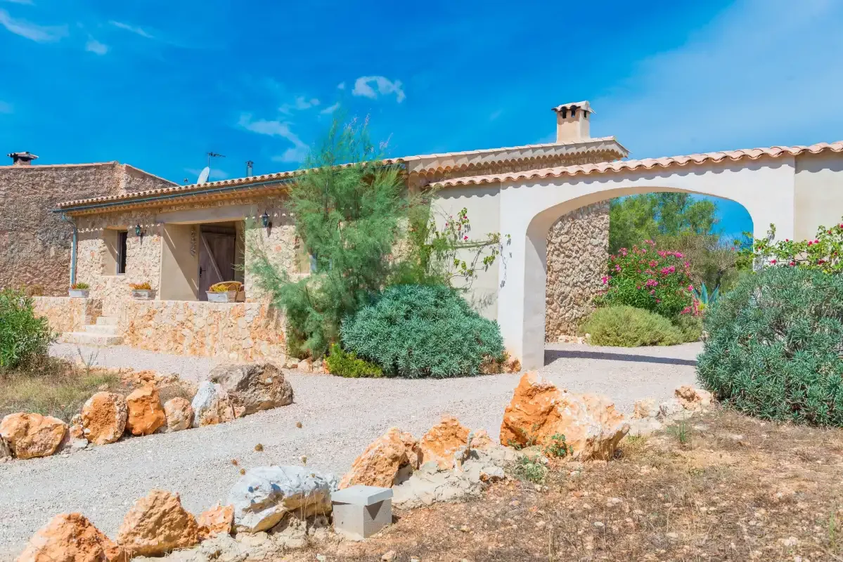 Una casa de piedra con una entrada arqueada está rodeada de rocas y arbustos floridos.