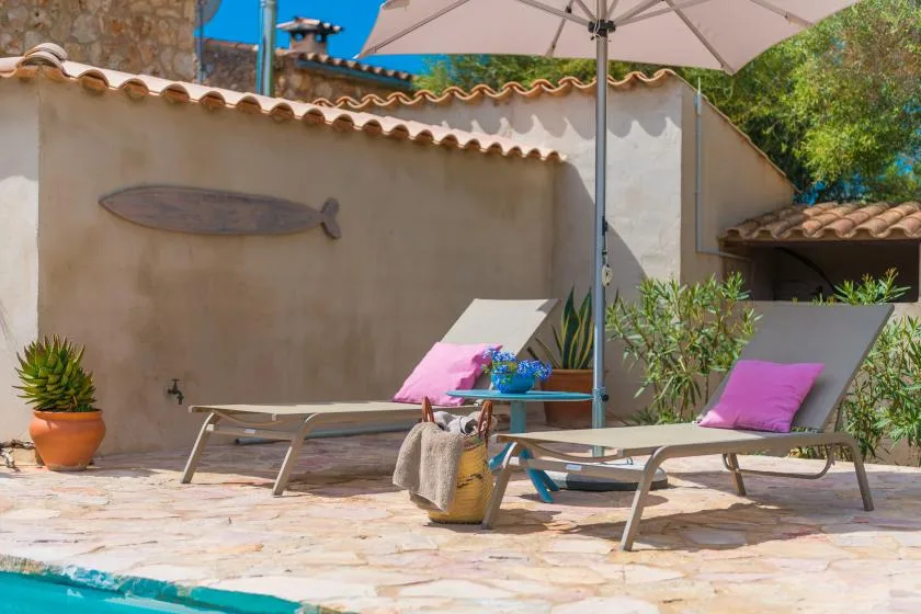 Un patio soleado tiene asientos reclinables bajo una sombrilla y cojines de colores.