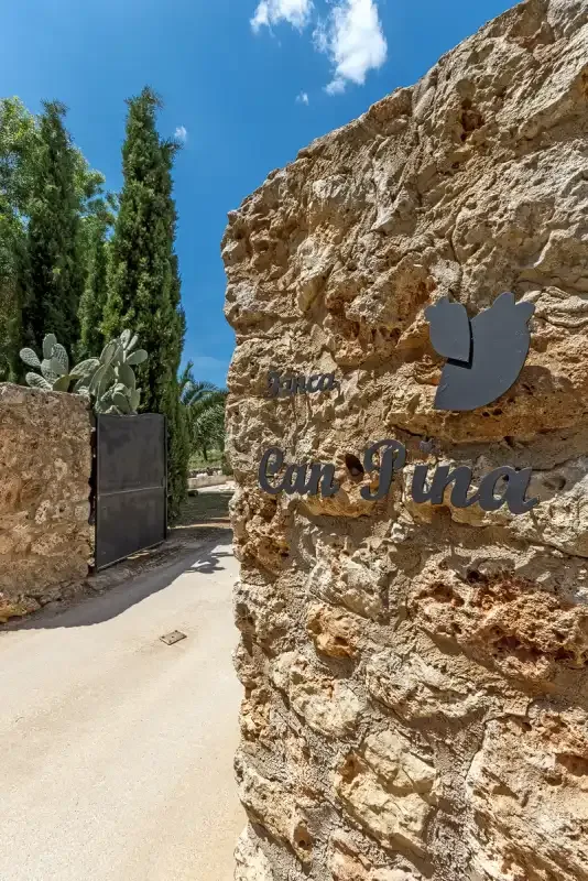 Un muro de piedra junto a un camino pavimentado, con un letrero y árboles.