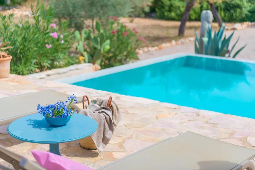 Una piscina junto a un patio de piedra con una mesa pequeña y plantas con flores.