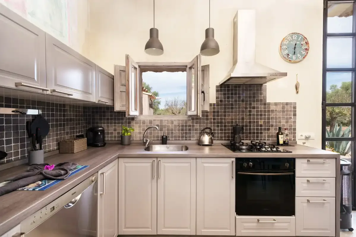 Una cocina luminosa con armarios blancos, azulejos oscuros y una ventana central.