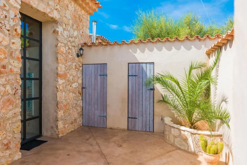 Un patio soleado con un muro de piedra, dos puertas de madera y palmeras en macetas.