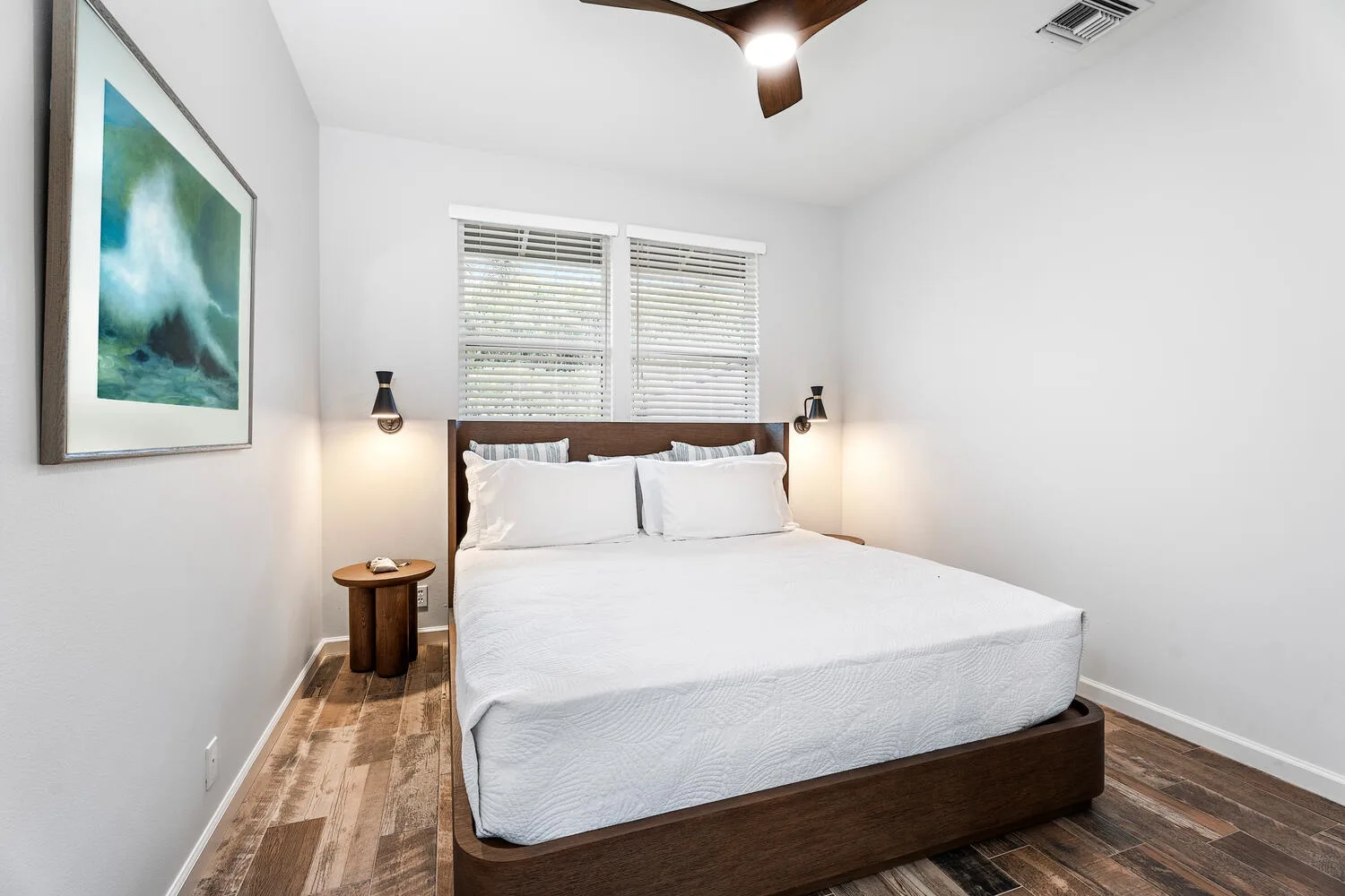 A bright bedroom with a bed beneath two windows with blinds.