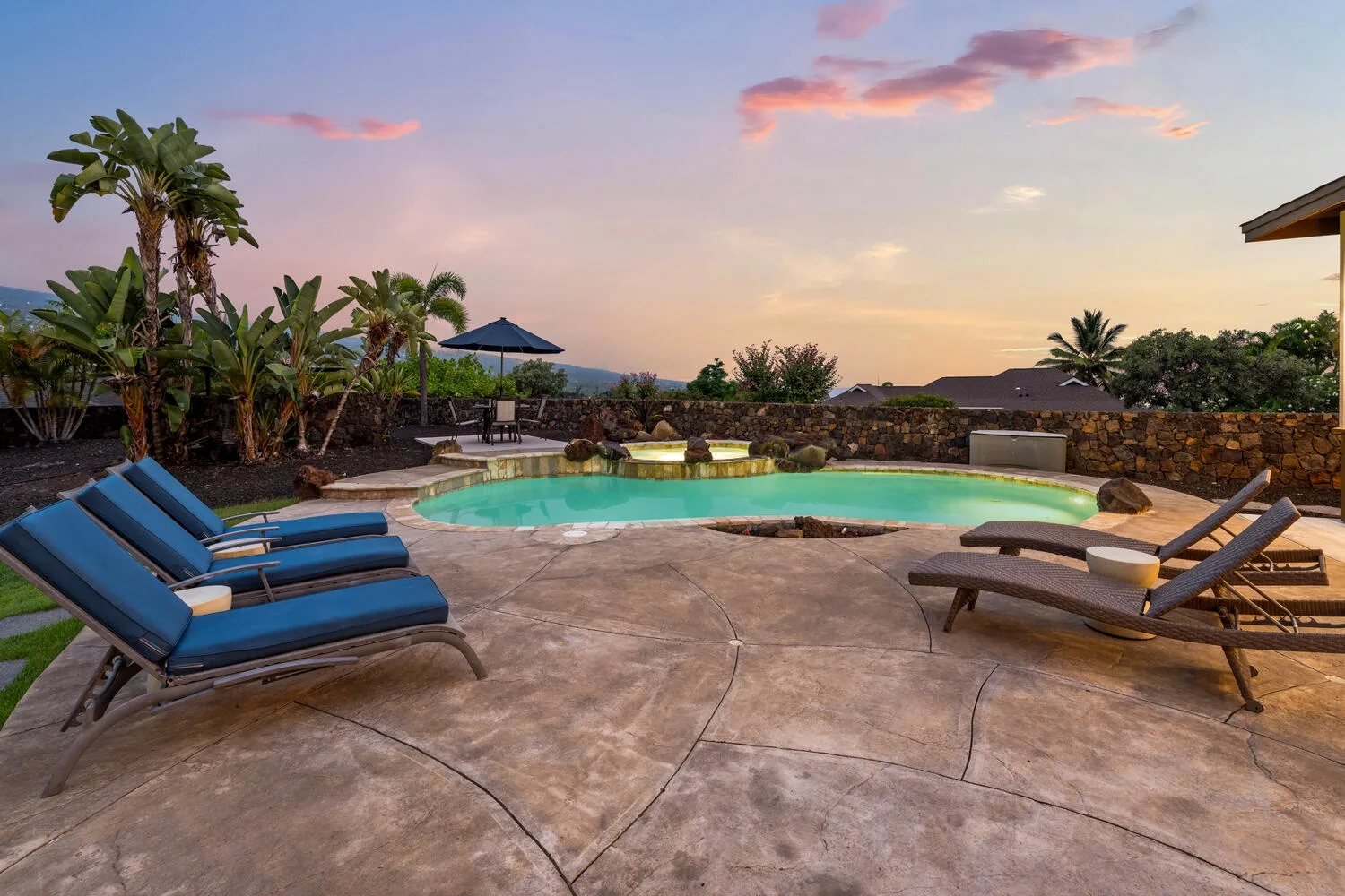 A swimming pool and surrounding lounge seating sit beneath a colorful sunset sky.