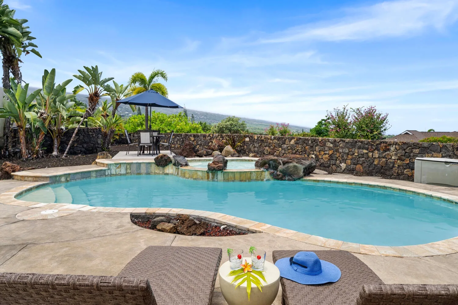 An outdoor swimming pool with lounge seating and tropical plants beside a stone wall.