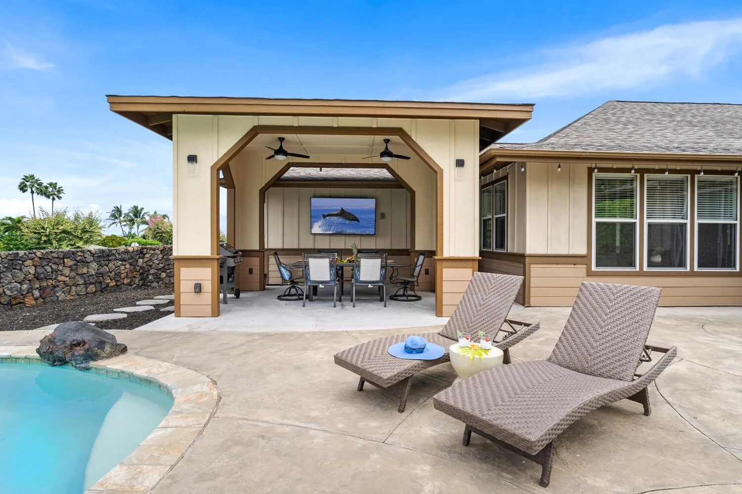 A covered outdoor seating area beside a swimming pool, with a dining table under the pavilion.