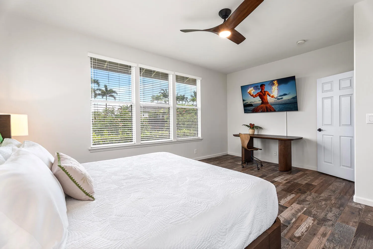 A bedroom with a bed, three windows, and a desk beneath a wall-mounted television.