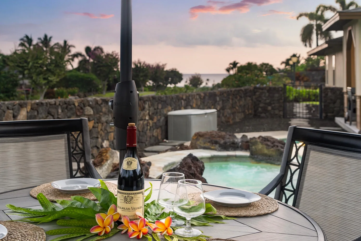 An outdoor dining table beside a pool, with tropical plants and a sunset view.