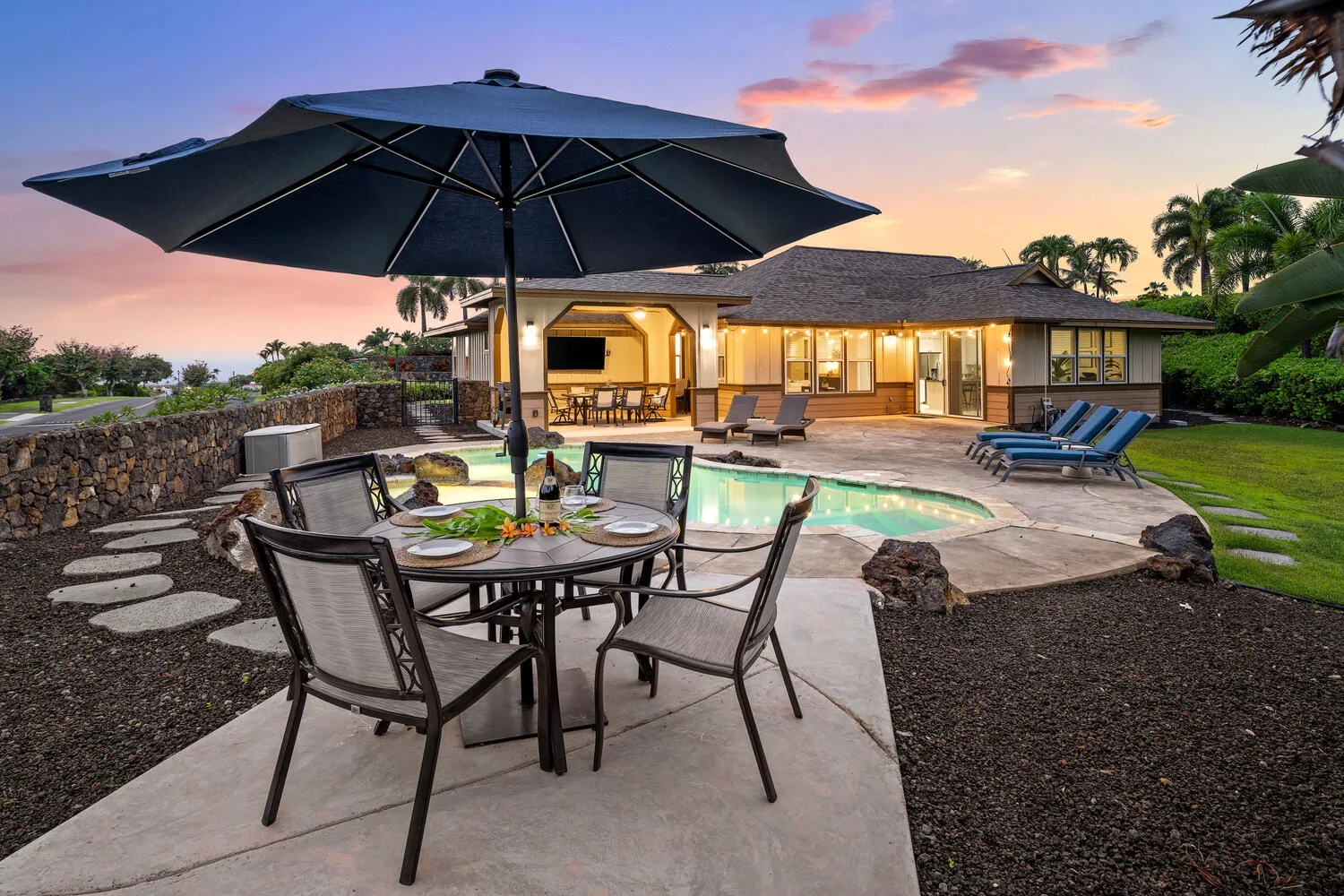 A patio dining area overlooks a swimming pool and a single-story house at sunset.