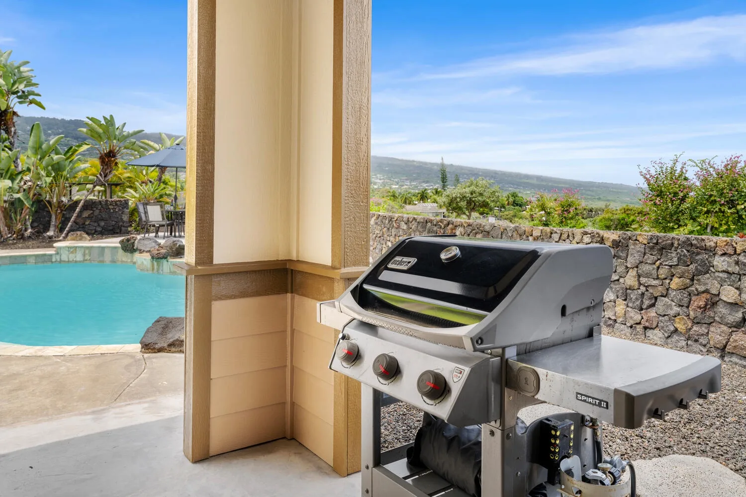 A barbecue grill sits beside a pool, with a stone wall and hills beyond.