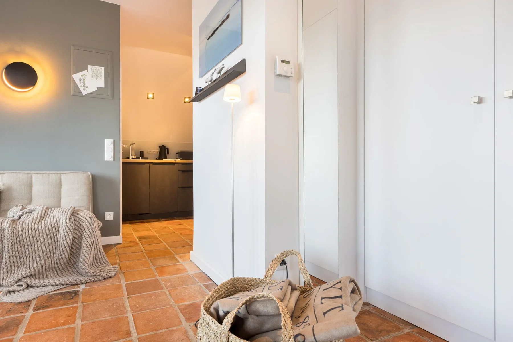 A living area with a sofa and an adjoining kitchen, featuring terracotta-colored floor tiles.