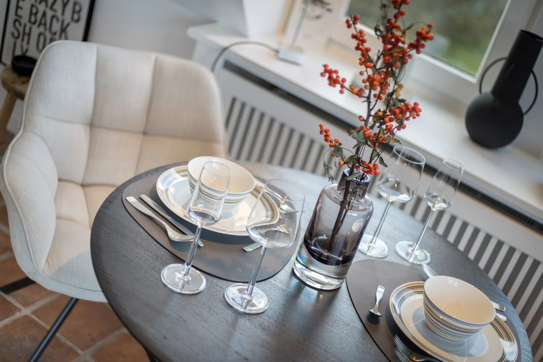 A dining table is set with dishes and glasses beside a window.