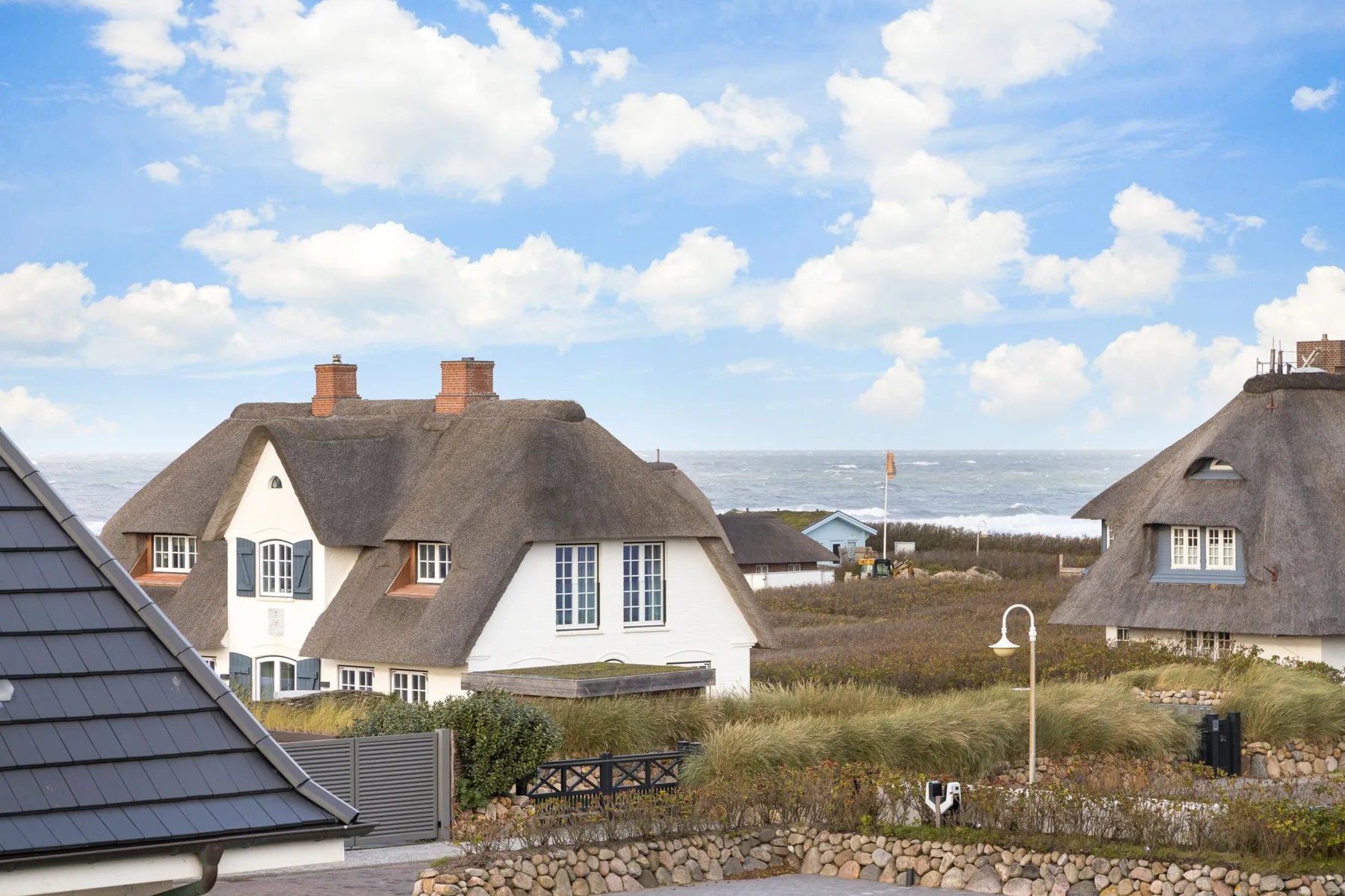 Thatched cottages sit among coastal dunes beneath a bright, cloud-filled sky.