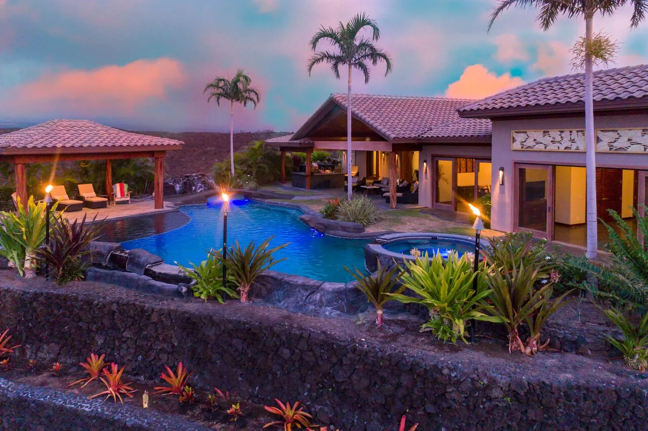 Un patio de estilo tropical con piscina iluminada y palmeras al atardecer.