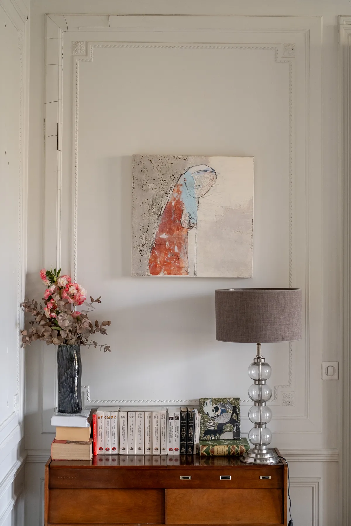Una habitación con paneles blancos, arte abstracto, libros, flores y una lámpara de mesa.
